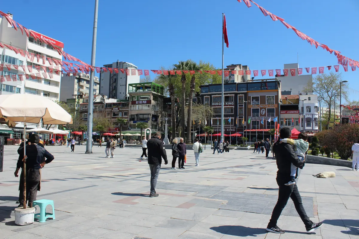 Bandırma Cumhuriyet Meydanı'nda güneşli bir günde yayalar, palmiyeler ve çevre işyerleri