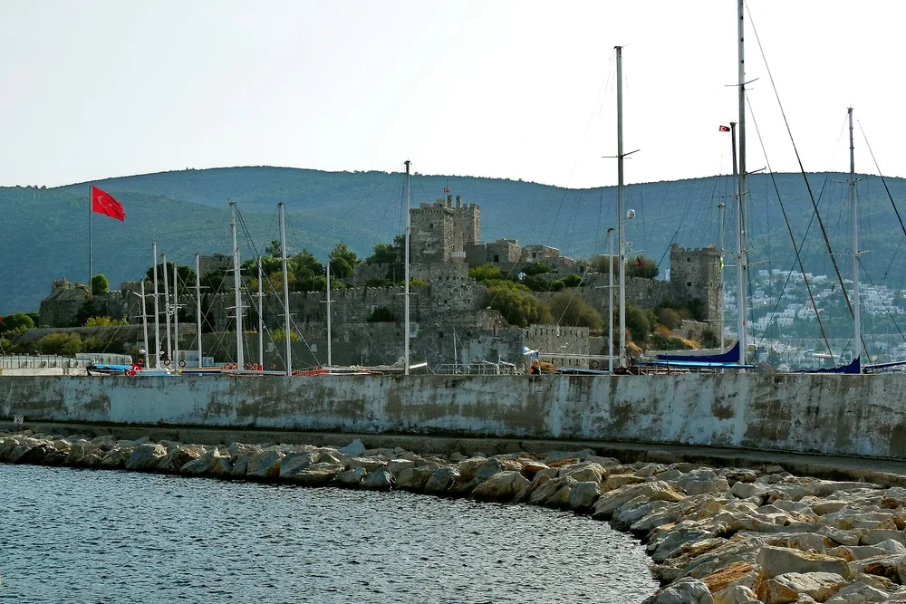 Bodrum Kalesi'nin mendirek tarafından görünümü, önde kayalık ve Türk bayrağı